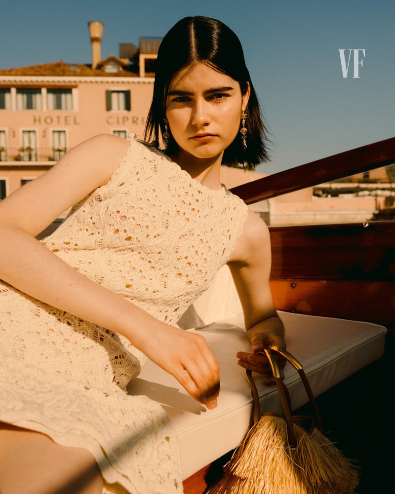 A fashionable woman in a sleeveless white dress poses on a boat with a building in the background, evoking elegance and style in Venice.