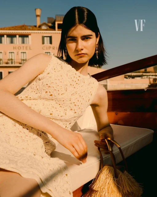 A fashionable woman in a sleeveless white dress poses on a boat with a building in the background, evoking elegance and style in Venice.