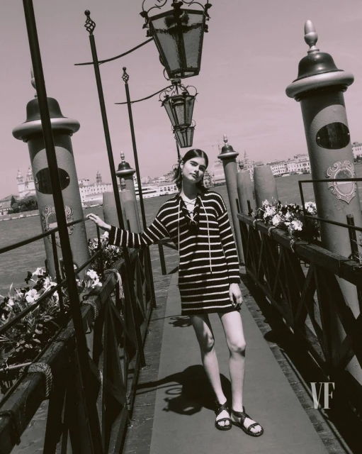 Woman in striped dress and sandals poses on a bridge in Venice, Italy, with historic buildings and a canal in the background.