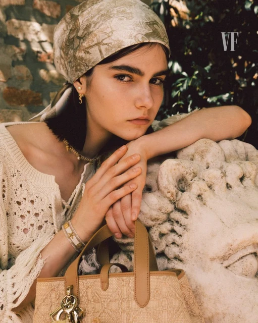 Young woman poses with handbag and headscarf near marble lion sculpture in an outdoor setting with natural light.  