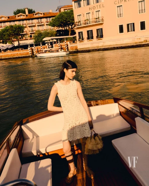 Woman in a white dress on a boat in Venice, Italy, with historic buildings and canal waters creating a scenic backdrop.