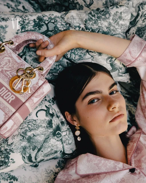 Woman in patterned pajamas lies on a toile-patterned blanket next to a Dior handbag with gold charms. Overhead shot, pink & green color palette.