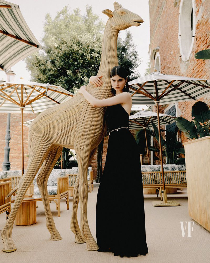 Woman in formal dress stands next to a giraffe statue outdoors on a patio with umbrellas, chairs, and a brick building.
