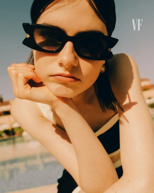 Young woman with black headband and sunglasses rests her chin on her hand with a pool and building in the background under a blue sky.