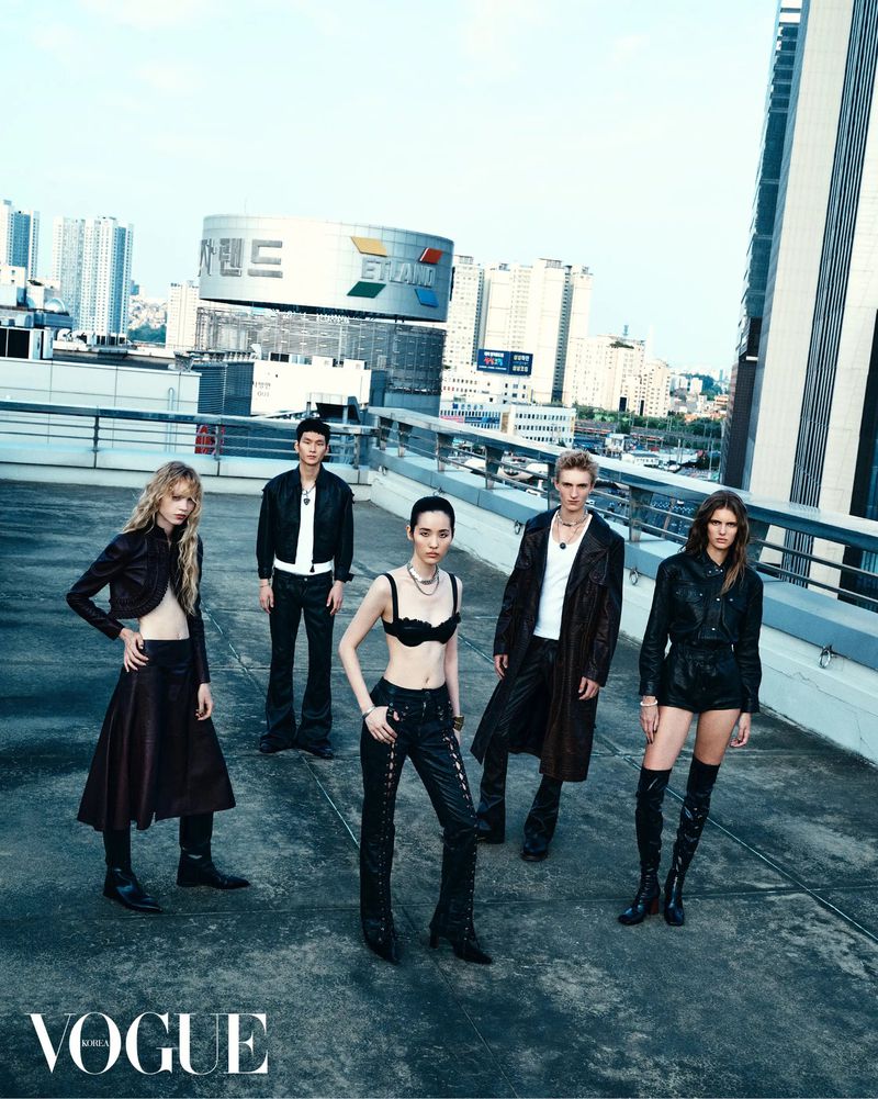 Fashion editorial shot of five models posing on a rooftop in black clothing, with city buildings in the background and the Vogue logo.