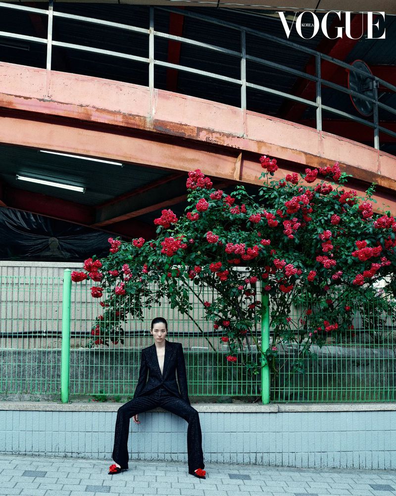 A fashion shot shows a woman in a black suit sitting on a ledge near red roses, wearing red heels against an urban backdrop, Vogue magazine.
