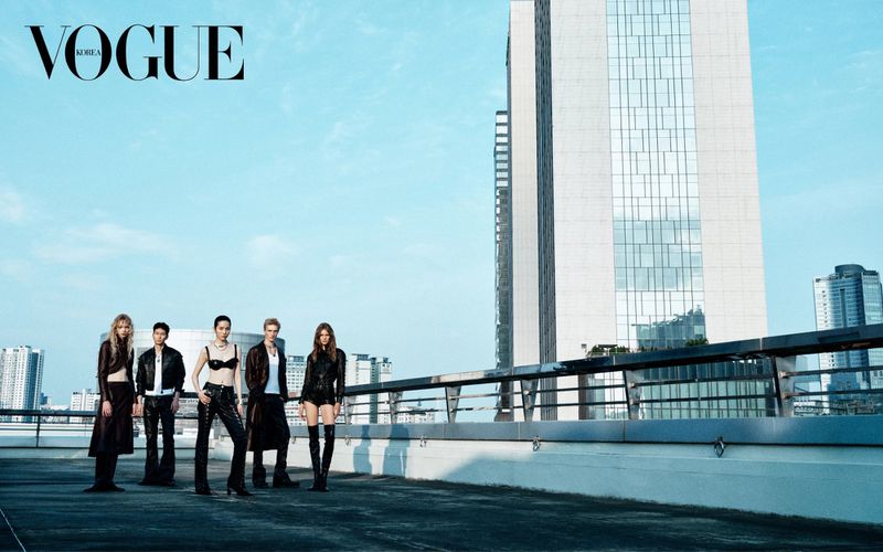 Fashion editorial featuring five models in edgy leather outfits on a rooftop with a city skyline and modern architecture backdrop.