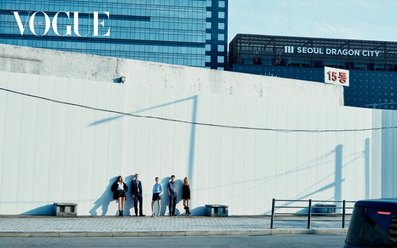 Editorial fashion shoot featuring five people standing near a white wall in an urban setting, with a modern building in the background and Vogue logo.