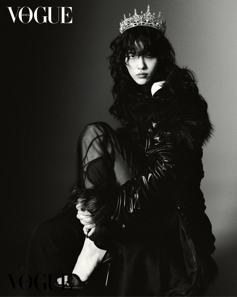 Model wearing a crystal tiara and dark textured coat seated against a gradient studio background