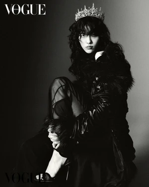 Model wearing a crystal tiara and dark textured coat seated against a gradient studio background