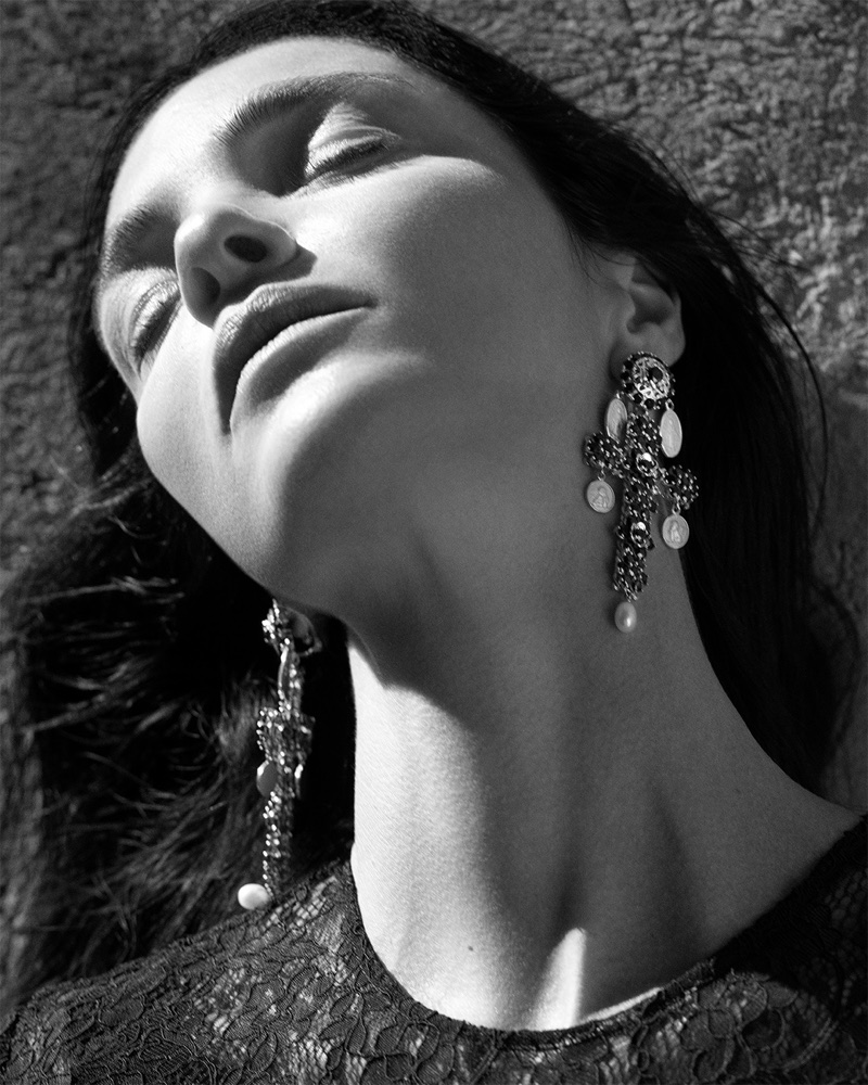 Close-up black and white portrait of a woman wearing ornate cross earrings and a lace top against a textured stone wall