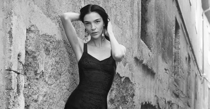 Mariacarla Boscono in a black slip dress posing against a weathered stone wall for Dolce & Gabbana DNA campaign