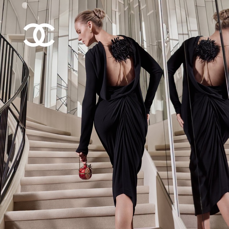 Model in a black cowl-back dress walking up a curved staircase, reflected in a large mirror.