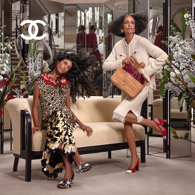 Two models in Chanel apparel posing in a luxurious salon with mirrored screens, floral arrangements, and a cream sofa.