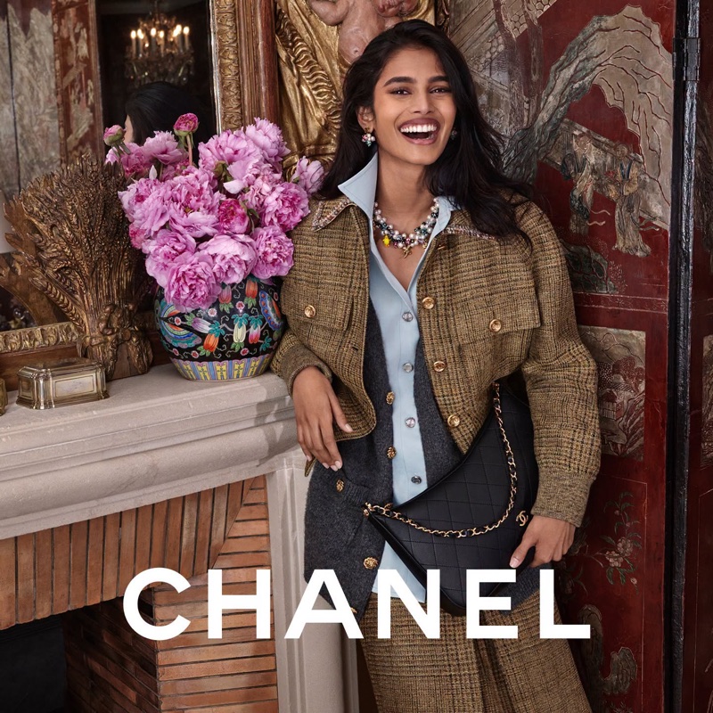 Smiling model leaning against a mantlepiece with pink flowers wearing a tweed jacket and carrying a black quilted handbag