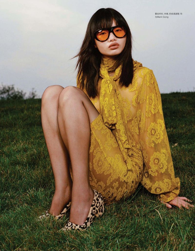 Woman in yellow lace dress and orange sunglasses sits on grass outdoors, wearing animal print heels in a fashion editorial shot.