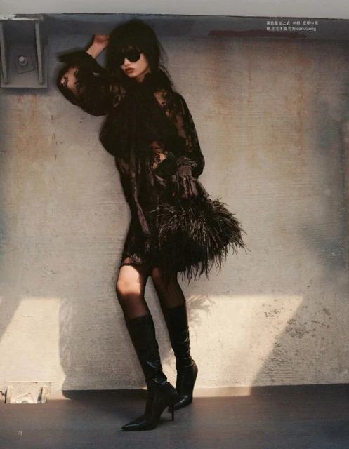 Model wearing dark sunglasses and black lace dress holding a feathered handbag against a textured concrete wall.