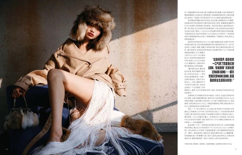Model wearing a faux fur hat and tan jacket holding a handbag seated on a blue sofa against a concrete wall