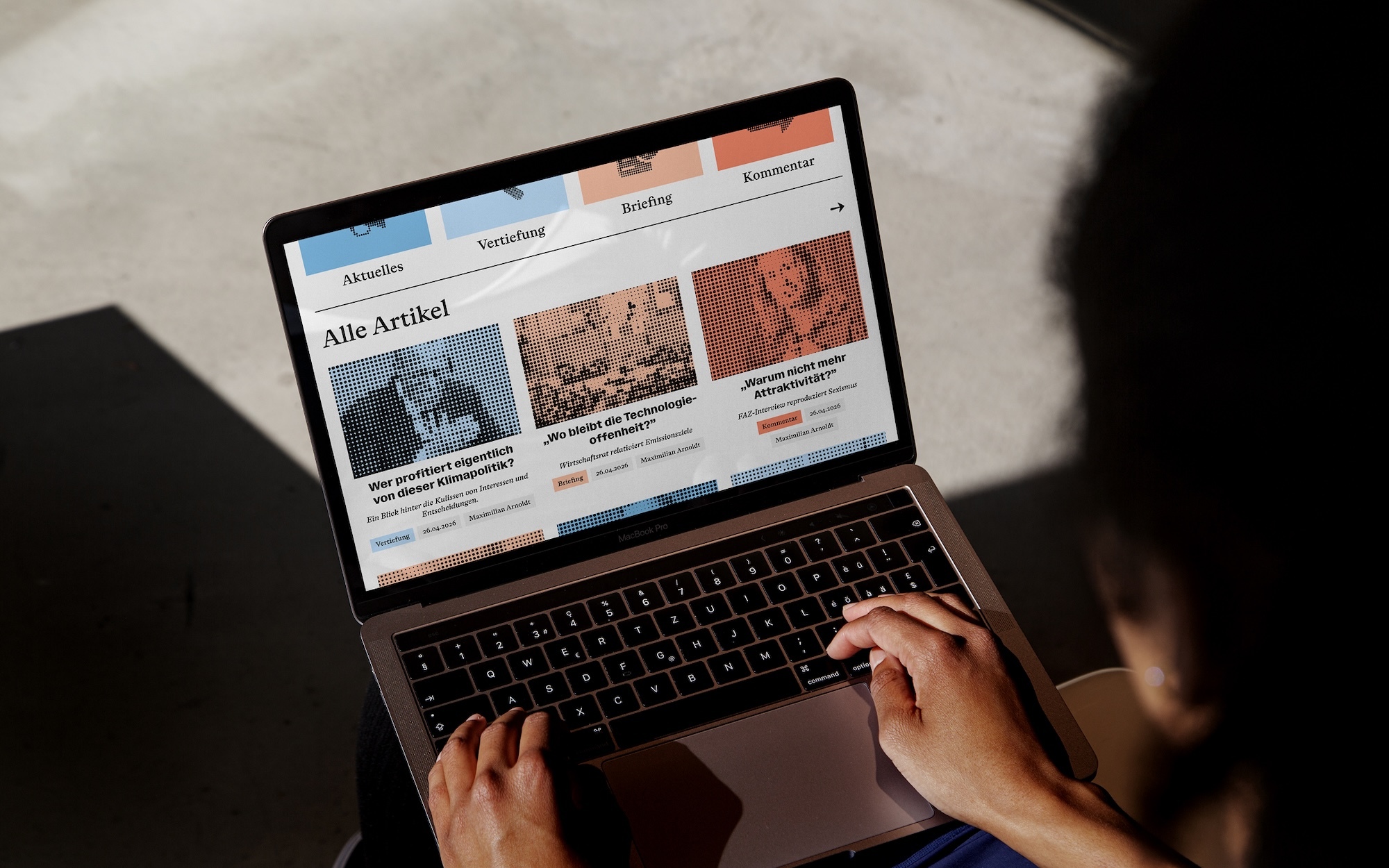 Close-up over-the-shoulder shot of hands typing on a laptop displaying a news website interface with orange halftone imagery.