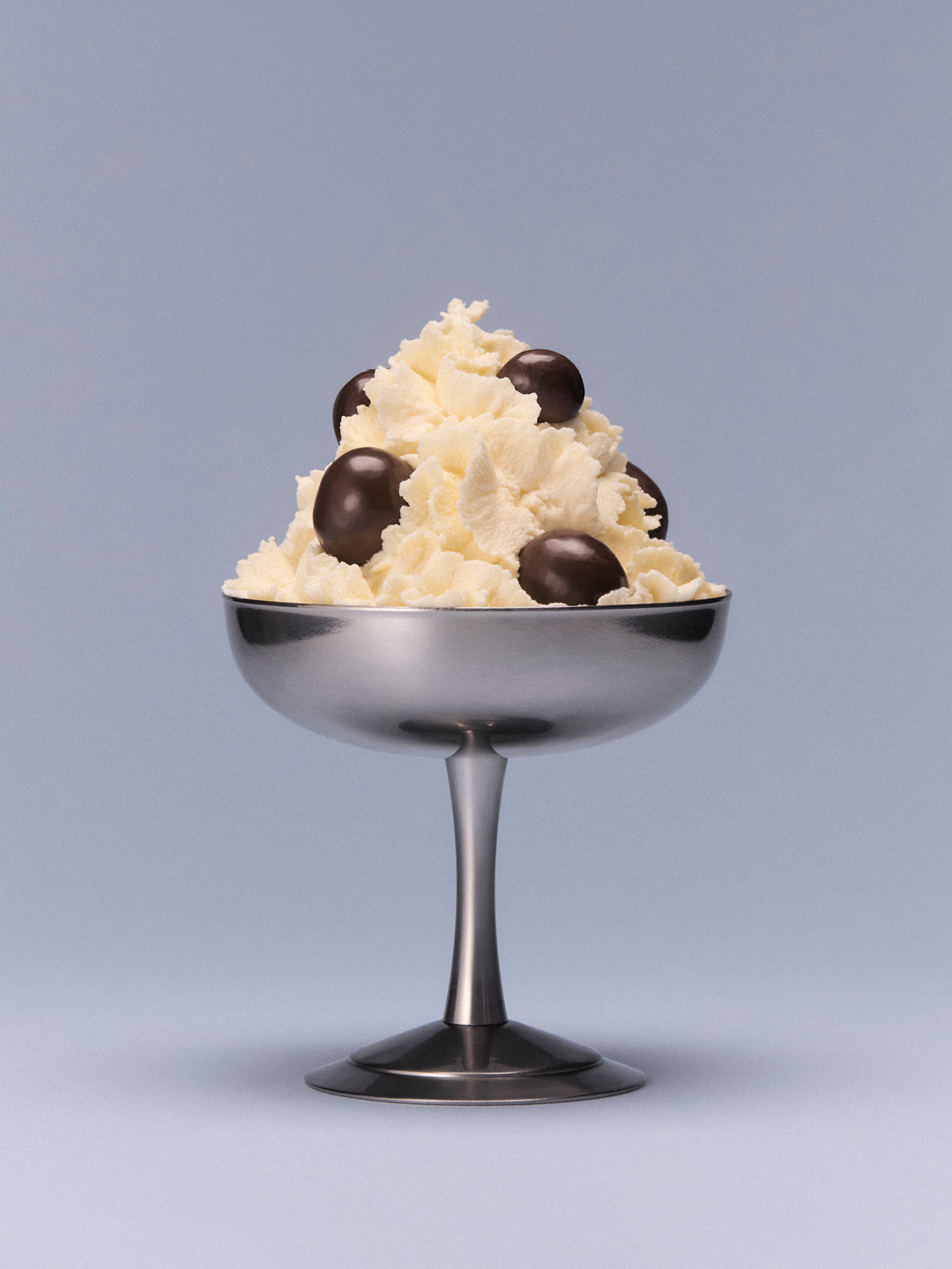 A metallic dessert goblet filled with whipped cream and dark chocolates on a light grey seamless background
