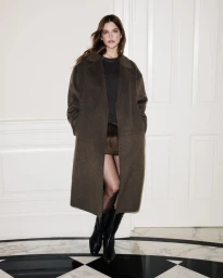 Barbara Palvin wearing a dark brown overcoat and black boots standing indoors against white panelled doors