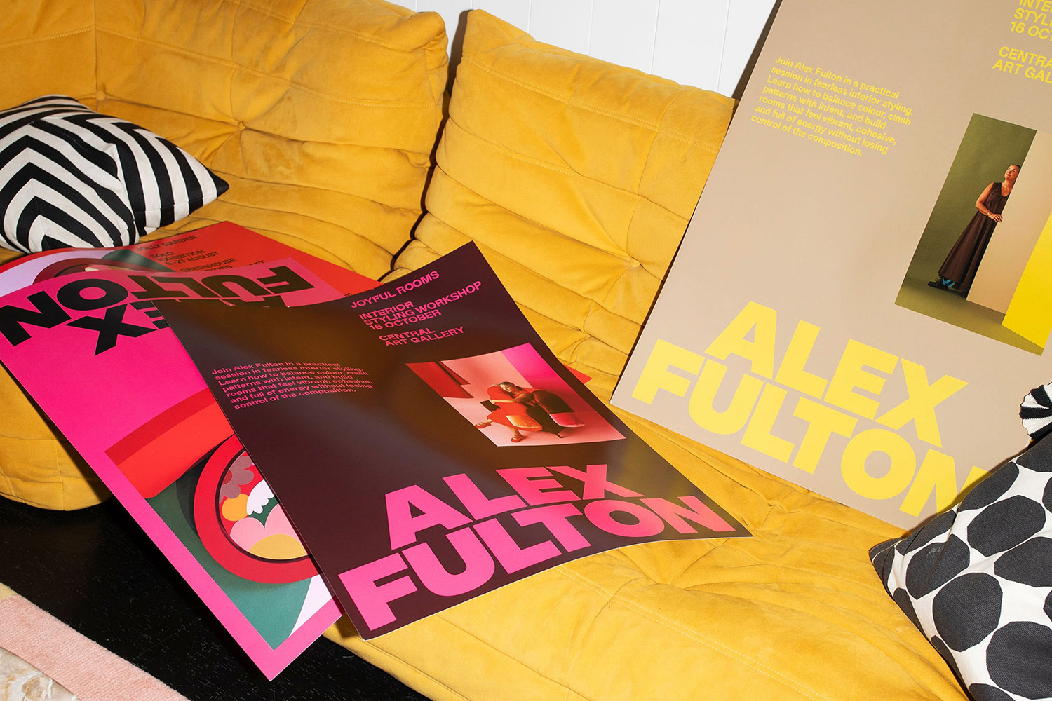 Vibrant posters for artist Alex Fulton displayed on a bright yellow quilted sofa with patterned pillows