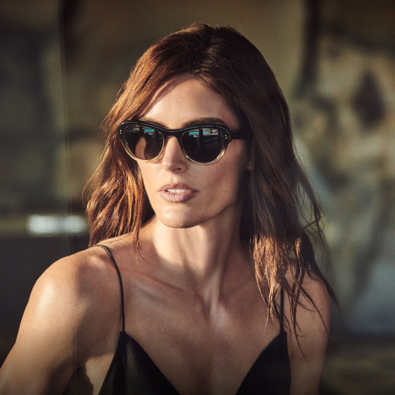 Model Hilary Rhoda wearing dark gradient sunglasses and a black top for Oliver Peoples