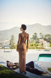 Model standing outdoors wearing a long brown dress near a pool with a reclining man and mountain views