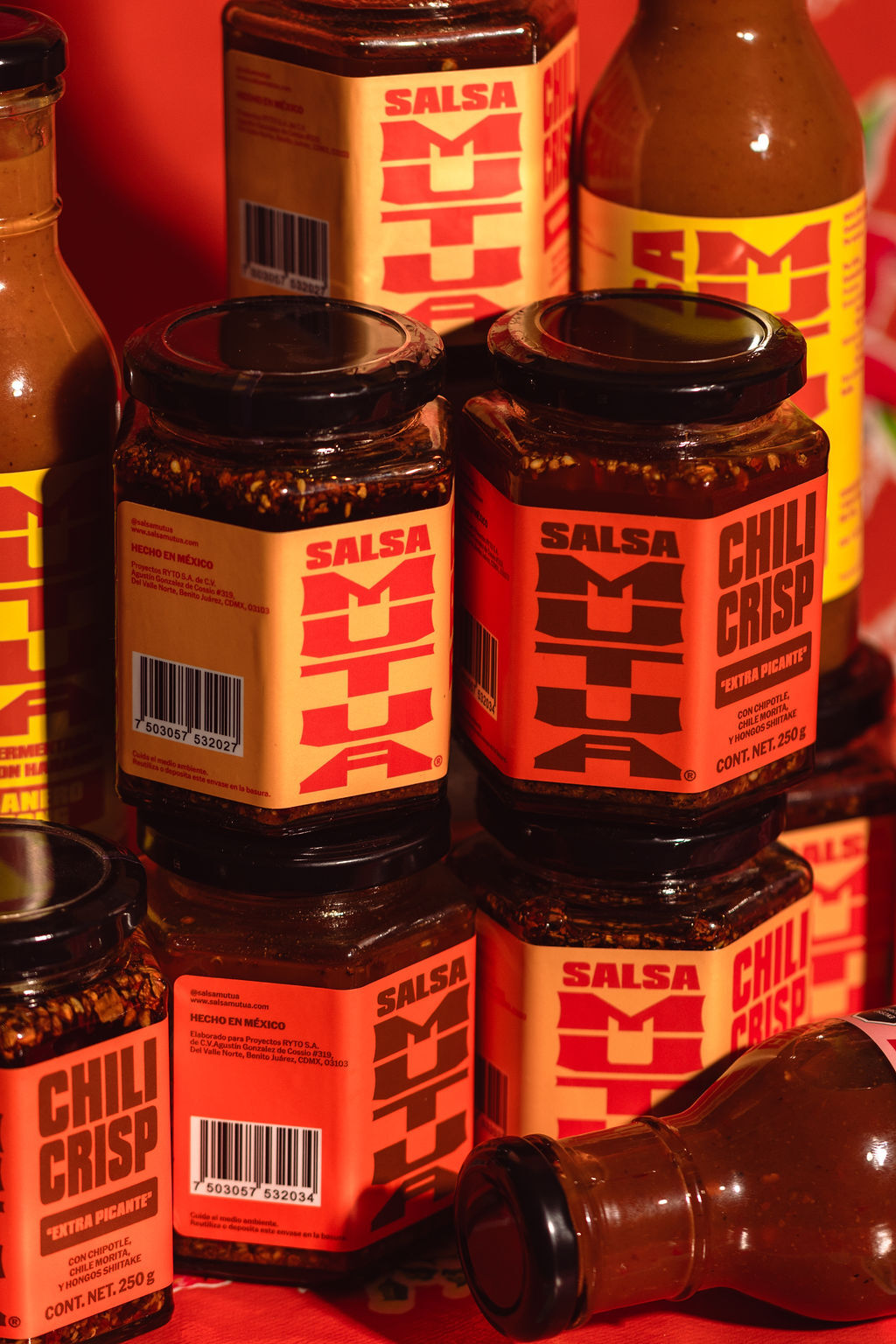 Close-up product shot of Salsa Mutua chili crisp jars and hot sauce bottles arranged on a red background.