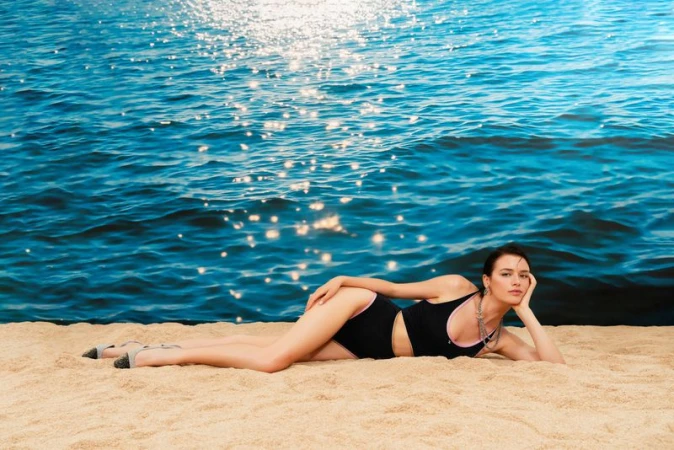 An adult female model lies on a sandy beach in a black swimsuit, posing in front of sparkling blue ocean water during daylight hours.