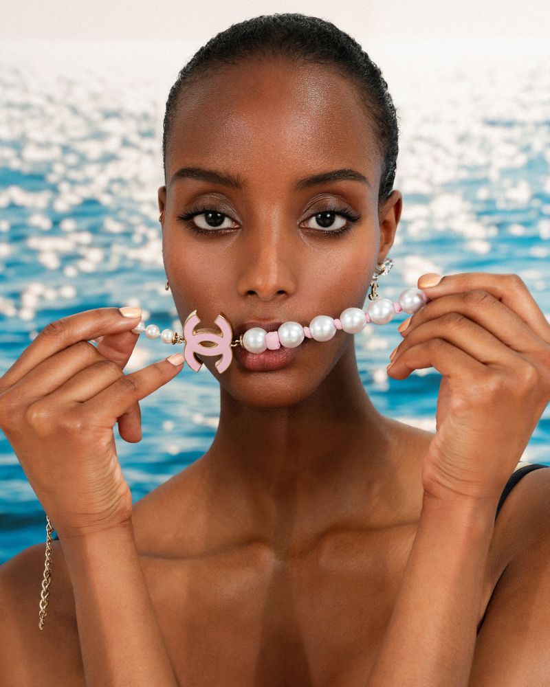 Portrait of a woman with dark skin holding a pearl necklace with a pink Chanel logo pendant, against a blurred ocean background.