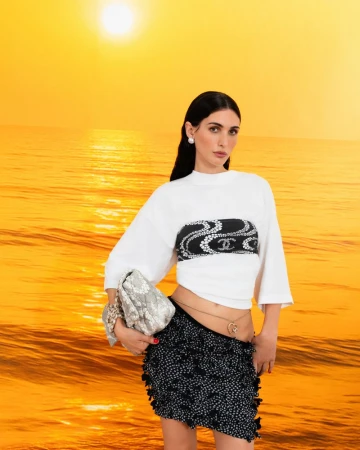 Woman in white crop top and black skirt poses against a golden sunset ocean backdrop, holding a light grey patterned purse.