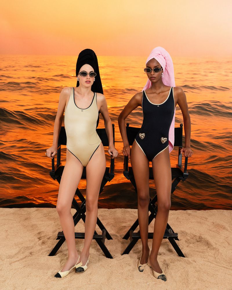 Two women in swimsuits and towels pose on director's chairs against an ocean sunset backdrop in a fashion photography studio setting.