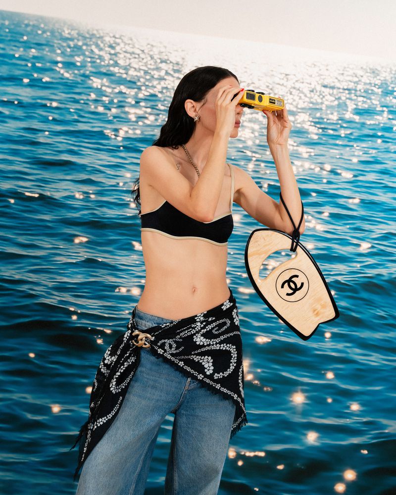 A woman stands in front of a blue sea backdrop, holding binoculars and a miniature surfboard; she's wearing jeans and a bikini top.