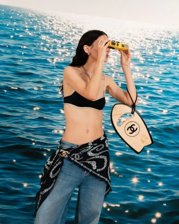 A woman stands in front of a blue sea backdrop, holding binoculars and a miniature surfboard; she's wearing jeans and a bikini top.