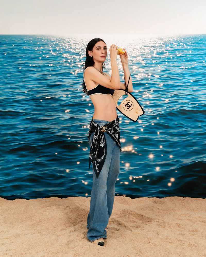 Woman in bikini top and jeans on a beach holding a yellow object, with ocean and horizon in the background on a sunny day.