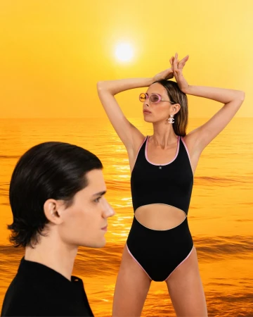 Two models pose against a warm beach sunset backdrop. One woman wears a black swimsuit, while the other is a man in a black jacket.