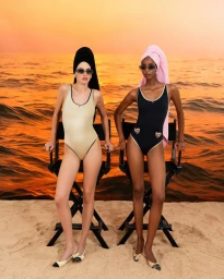 Two female models in swimsuits and towels on their heads posing in director chairs against a sunset beach backdrop for Grazia Italy.