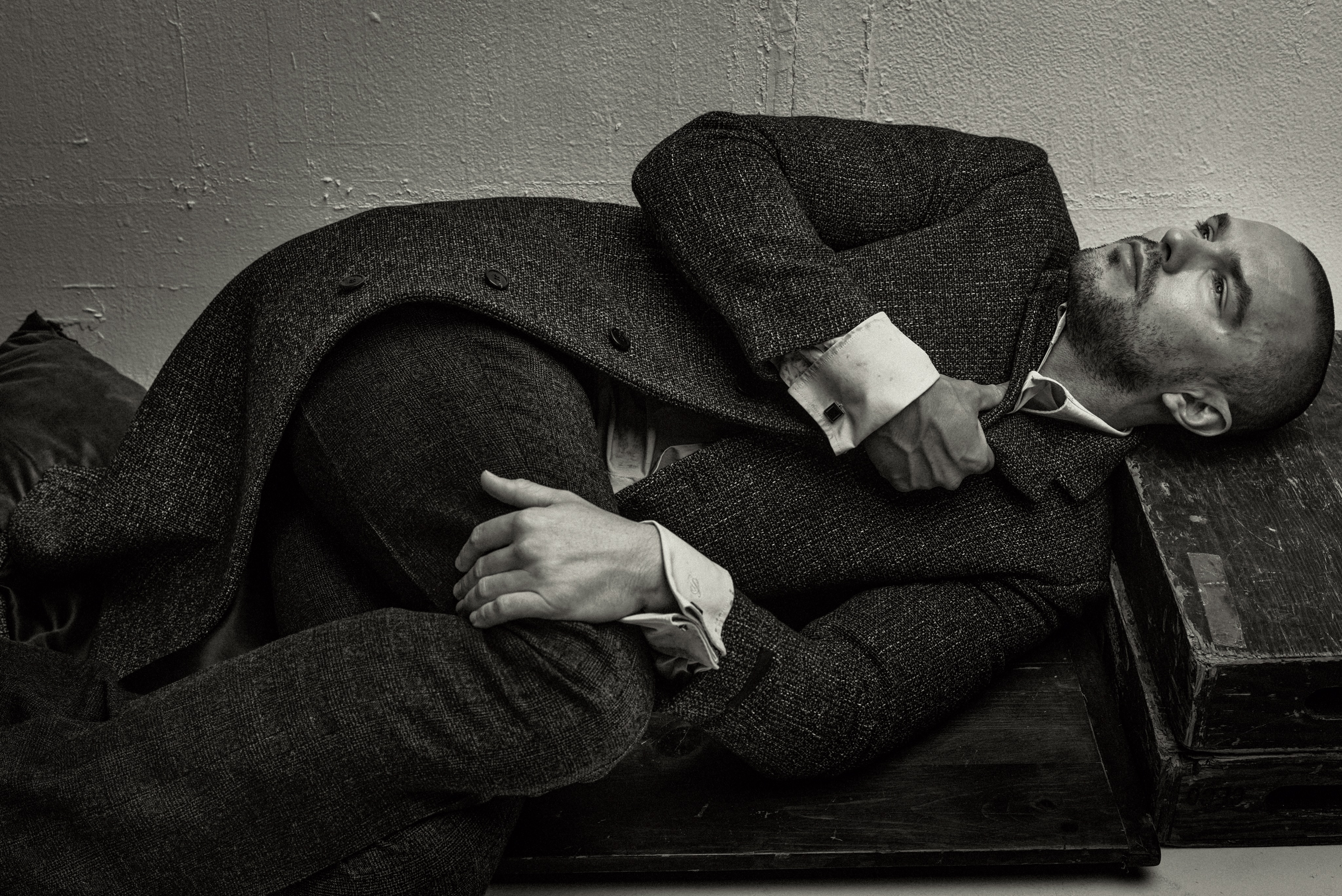 Nicholas Hoult wearing a dark tweed coat and suit lying down in a monochrome portrait for ICON America