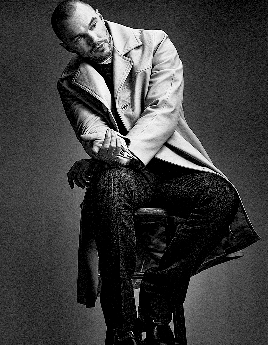 Nicholas Hoult seated on a stool wearing a long coat and dark trousers in a studio portrait by Craig McDean.