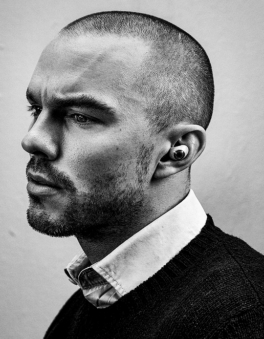Monochrome portrait of Nicholas Hoult in profile wearing a collared shirt and dark sweater against a plain background