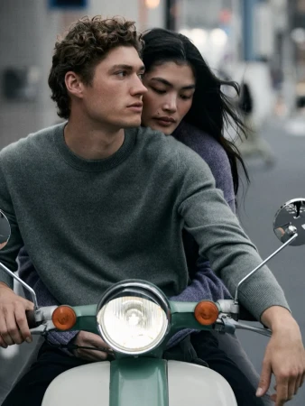 Man with curly hair driving a scooter with a woman embracing him from behind on a city street