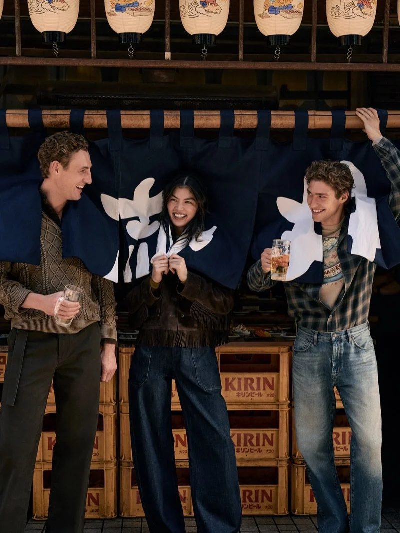 Three smiling models posing under a blue noren curtain with beer crates in a street-side scene for Banana Republic September 2026