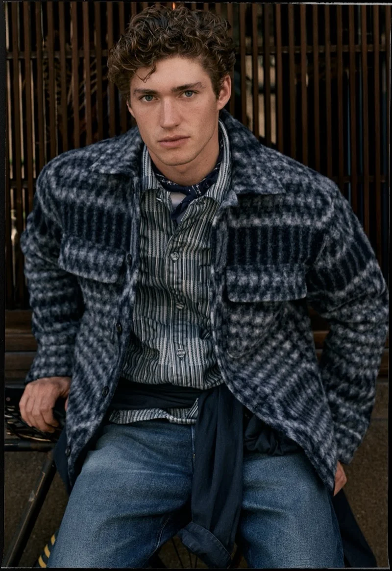 Young man with curly hair wearing a plaid overshirt, striped shirt, and blue jeans against a slated wooden background