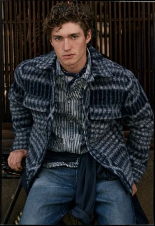 Young man with curly hair wearing a plaid overshirt, striped shirt, and blue jeans against a slated wooden background
