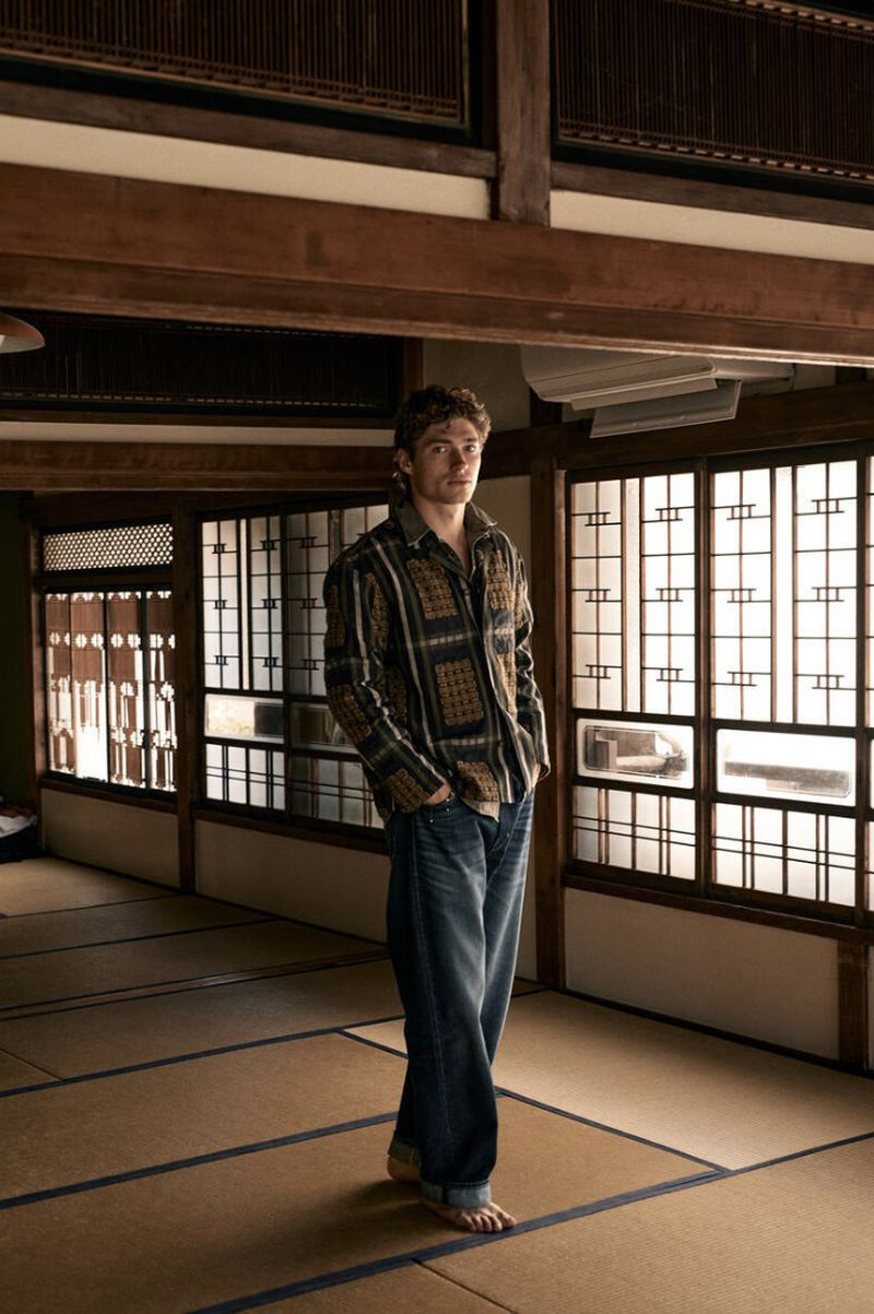 A man wearing a patterned long-sleeve shirt and jeans standing barefoot on tatami mats inside a traditional interior.