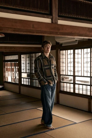 A man wearing a patterned long-sleeve shirt and jeans standing barefoot on tatami mats inside a traditional interior.
