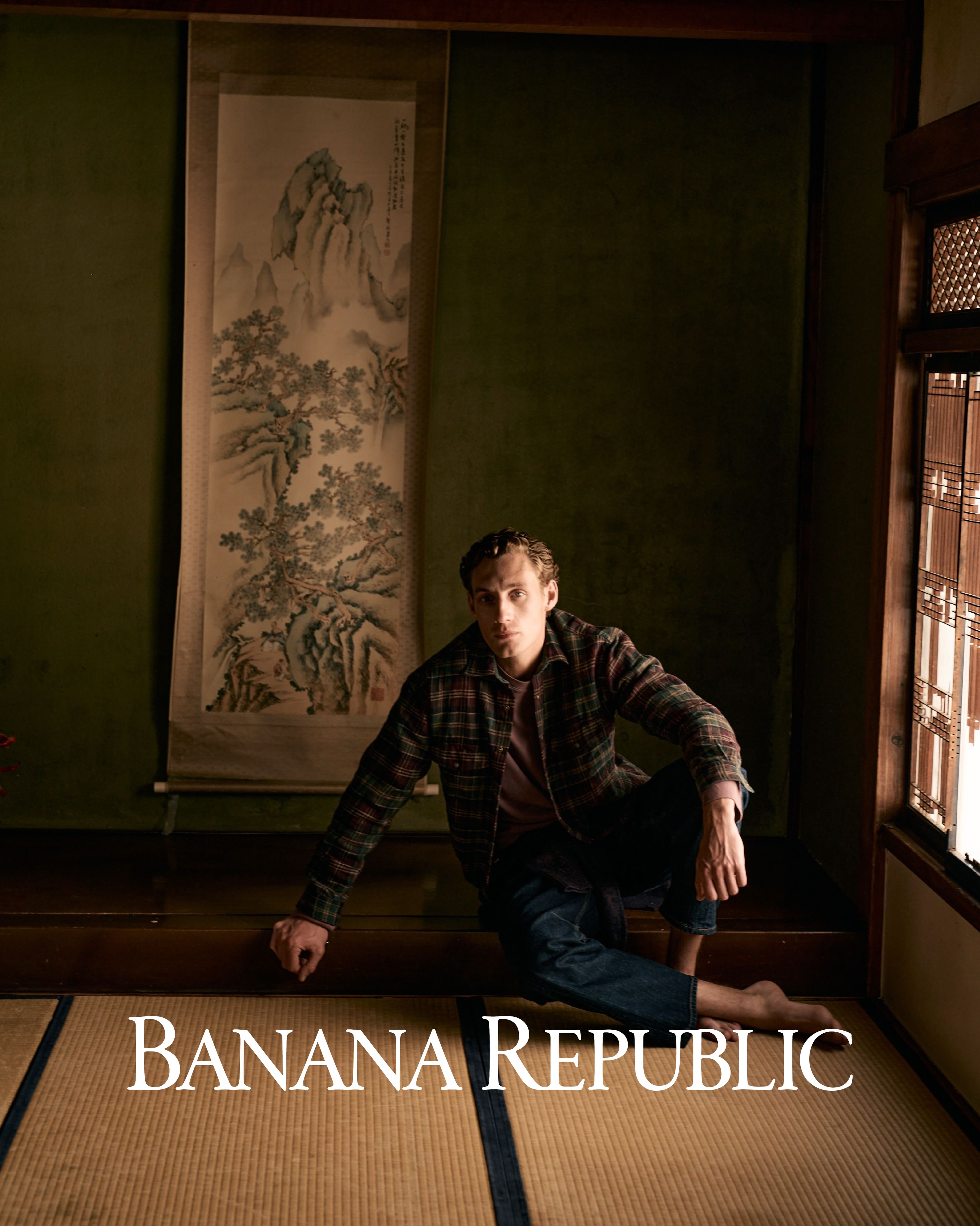 Man seated on tatami mat wearing a plaid jacket and dark trousers inside a traditional interior with a hanging landscape scroll.