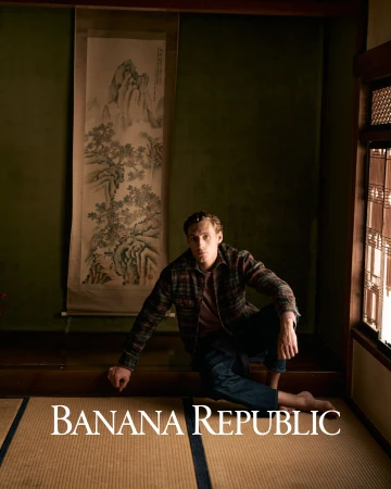 Man seated on tatami mat wearing a plaid jacket and dark trousers inside a traditional interior with a hanging landscape scroll.
