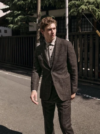 A man wearing a brown suit and patterned shirt standing on a paved street outdoors.